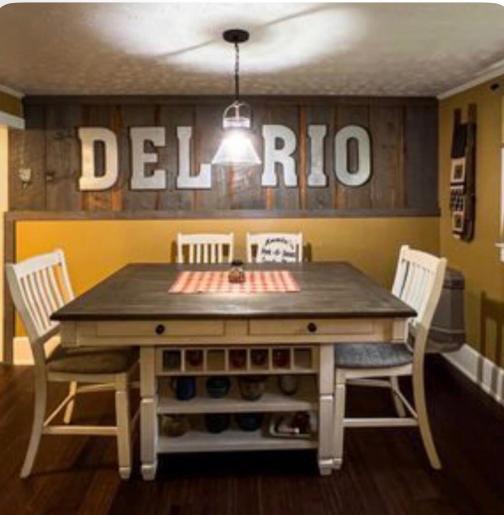 Farmhouse dining room with a long wood table, white chairs, and a reclaimed-wood DEL RIO sign on the wall
